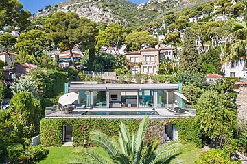 Aerial view of a modern hillside villa with an infinity pool, surrounded by lush trees and traditional Mediterranean homes.