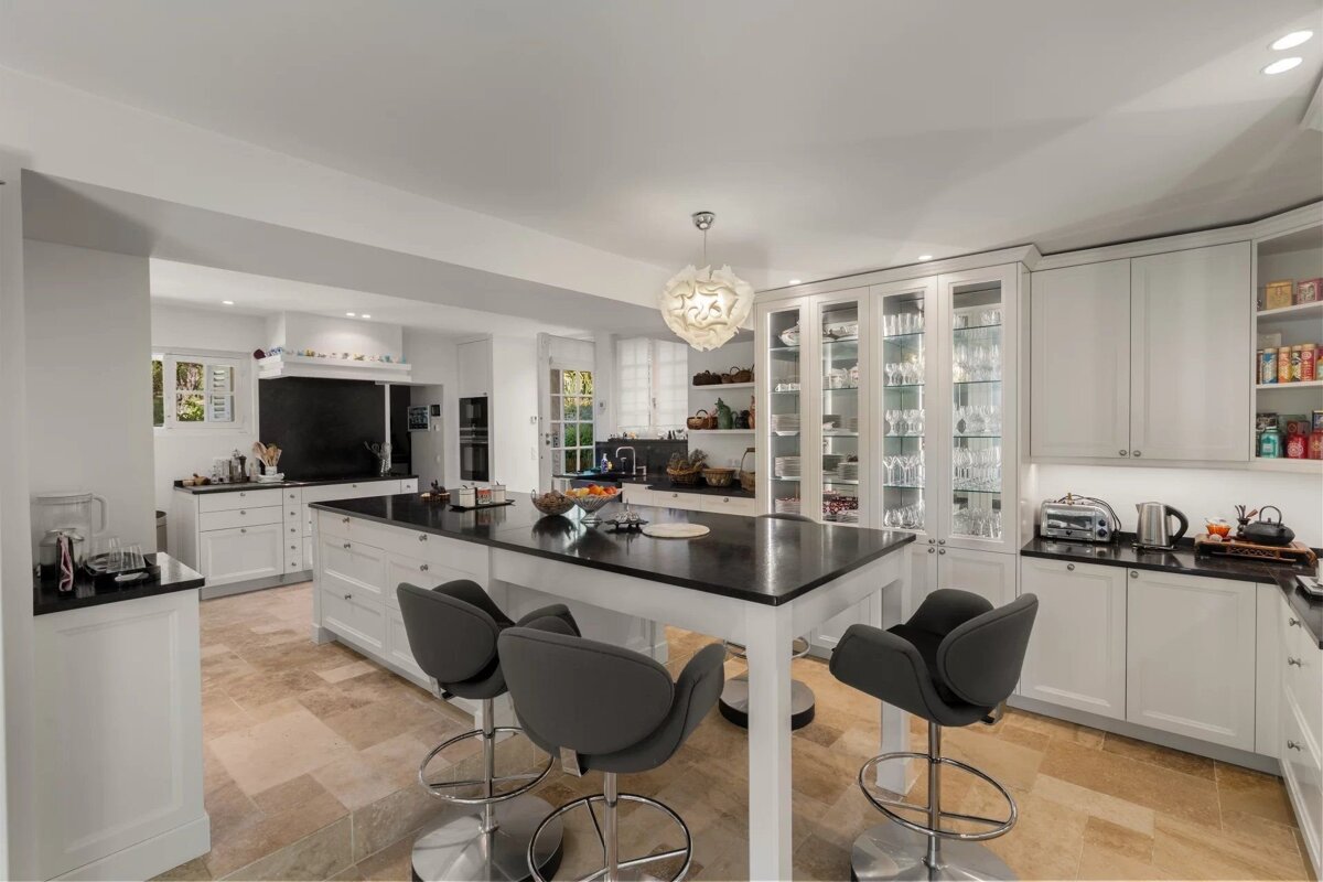 A spacious, modern white kitchen featuring a large black island with grey bar stools, tiled floor, and white cabinets, some with glass fronts.