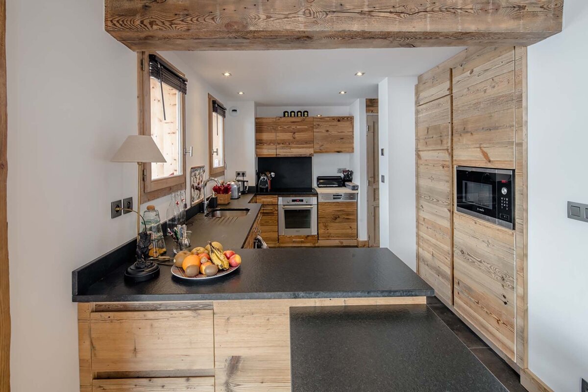 Chalet-style kitchen with rustic wood cabinets, dark countertops, integrated appliances, and a fruit bowl on the island, framed by a large wooden beam.