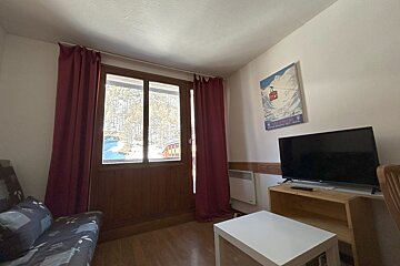 A cozy room with a large window overlooking a snowy landscape, red curtains, a sofa, TV, coffee table, and a ski poster.