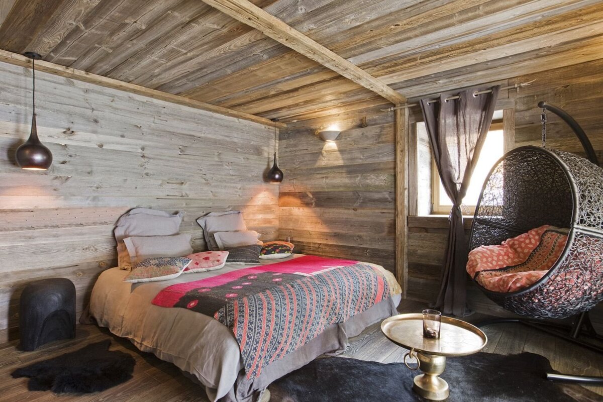 A cozy, rustic wood-paneled bedroom with a colorful bed and a hanging egg chair. Warm lighting creates an inviting, chalet-style ambiance.