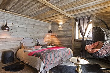 A cozy, rustic wood-paneled bedroom with a colorful bed and a hanging egg chair. Warm lighting creates an inviting, chalet-style ambiance.