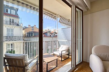 A sunny balcony with outdoor seating and city buildings view, seen through open doors from an indoor room with a cream armchair.