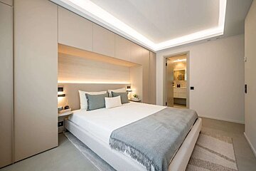 A modern, neutral-toned bedroom with built-in storage, soft ambient lighting, a white bed, and a partially visible en-suite bathroom.