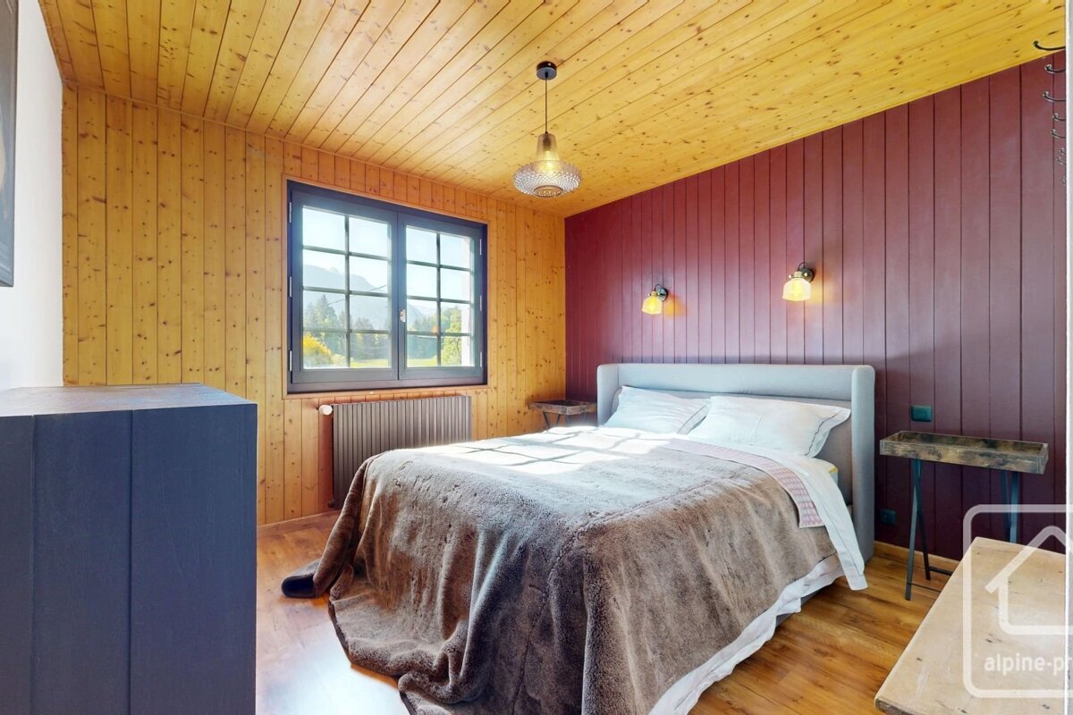 Cozy bedroom featuring warm wood walls and ceiling, a dark red accent wall, and a bed. A large window offers stunning mountain views.