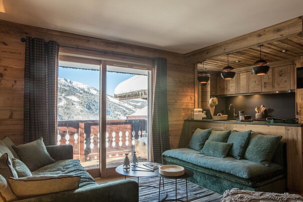 Cozy wooden chalet living room and kitchen with plush sofas, overlooking a stunning snowy mountain landscape through a large window.