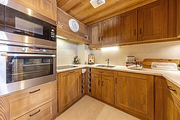 A cozy, modern L-shaped kitchen with warm wooden cabinets, stainless steel appliances (oven, microwave), a sink, and white countertops under bright lighting.