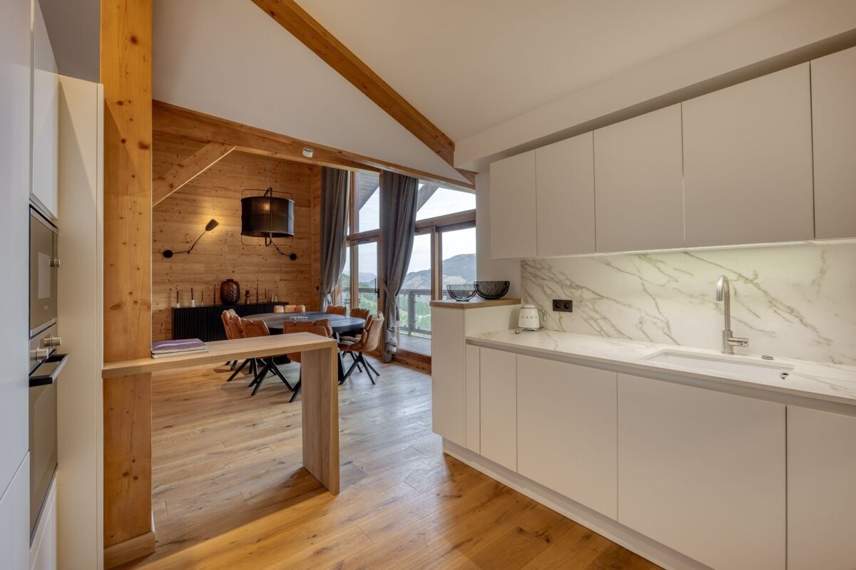 Modern white kitchen with marble backsplash, open to a cozy, wood-paneled dining area featuring large windows and mountain views.