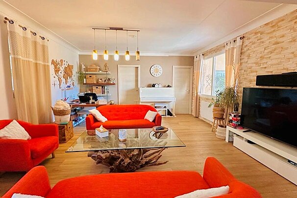 A colorful, cozy living room with bright orange sofas, a glass table, world map, TV, and a white piano, featuring modern and rustic touches.