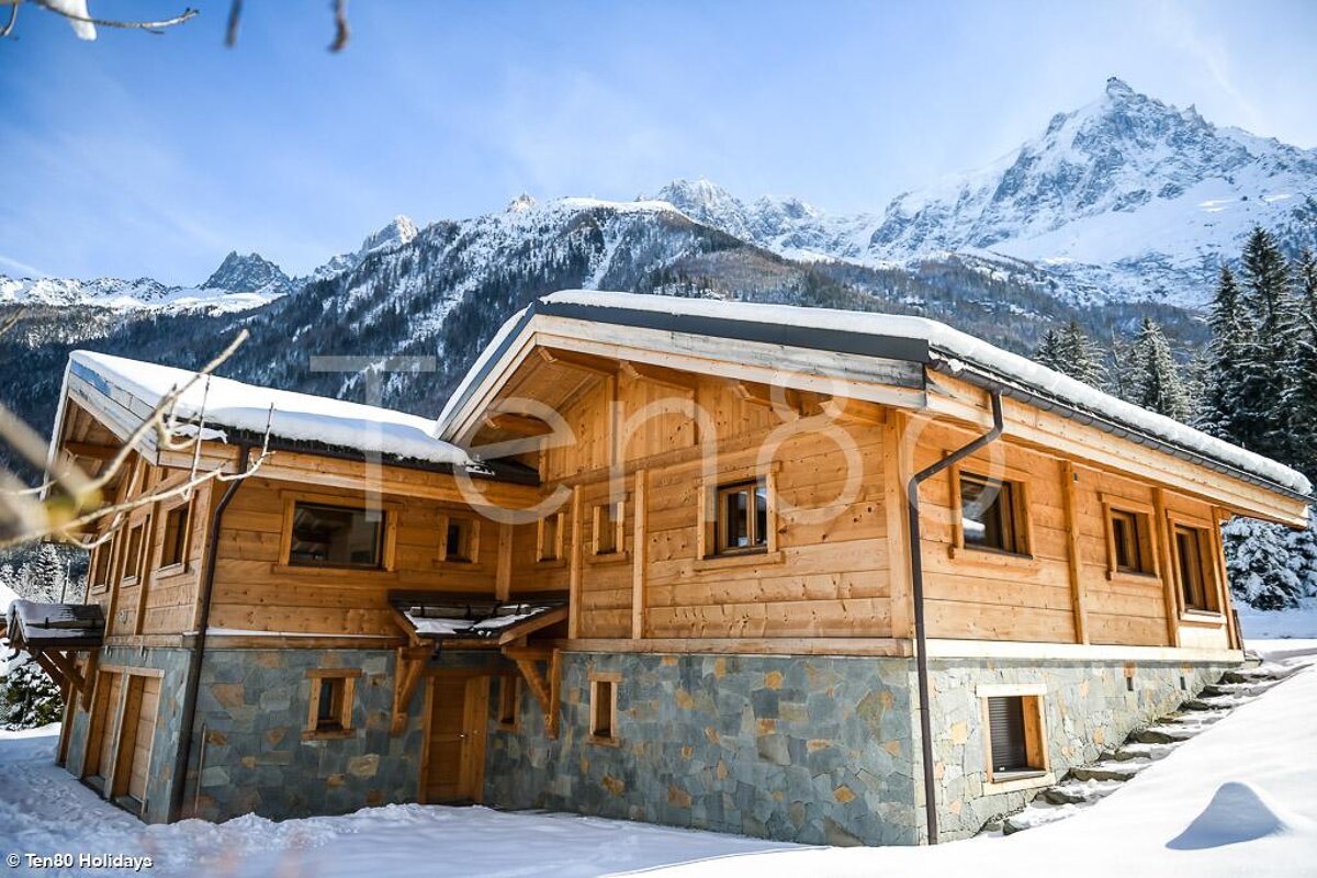 A large wooden house with snow on the roof and mountains in the background