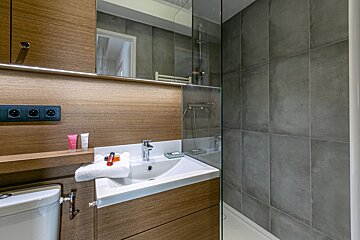 A modern bathroom with a wooden vanity, sink, and toilet next to a grey-tiled walk-in shower with a glass screen.