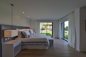 Modern bedroom with a large bed, sleek hanging lamps, wooden floor, and floor-to-ceiling windows opening to a lush green lawn.