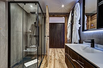 A modern bathroom features a glass shower, textured gray walls, and light wood flooring. A black vanity, mirror with LED light, and white robes complete the stylish look.