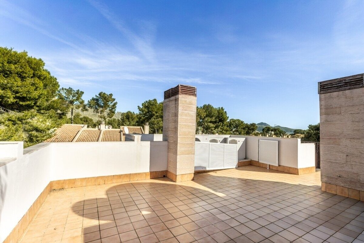 Sunny rooftop terrace with tiled floor, white walls, and a chimney. Green trees, distant hills, and other rooftops under a clear blue sky.