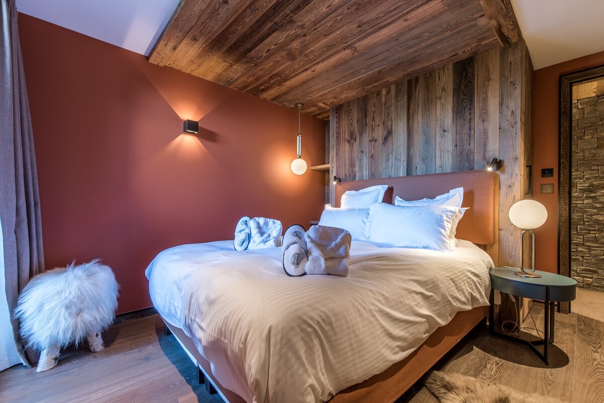 A cozy, modern alpine bedroom featuring wood-paneled accents, terracotta walls, white bedding, and warm ambient lighting.