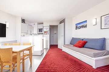 A bright, open-plan studio with a dining area, white kitchen, white sofa bed, and a striking red geometric rug. Ski-themed art adorns the walls.