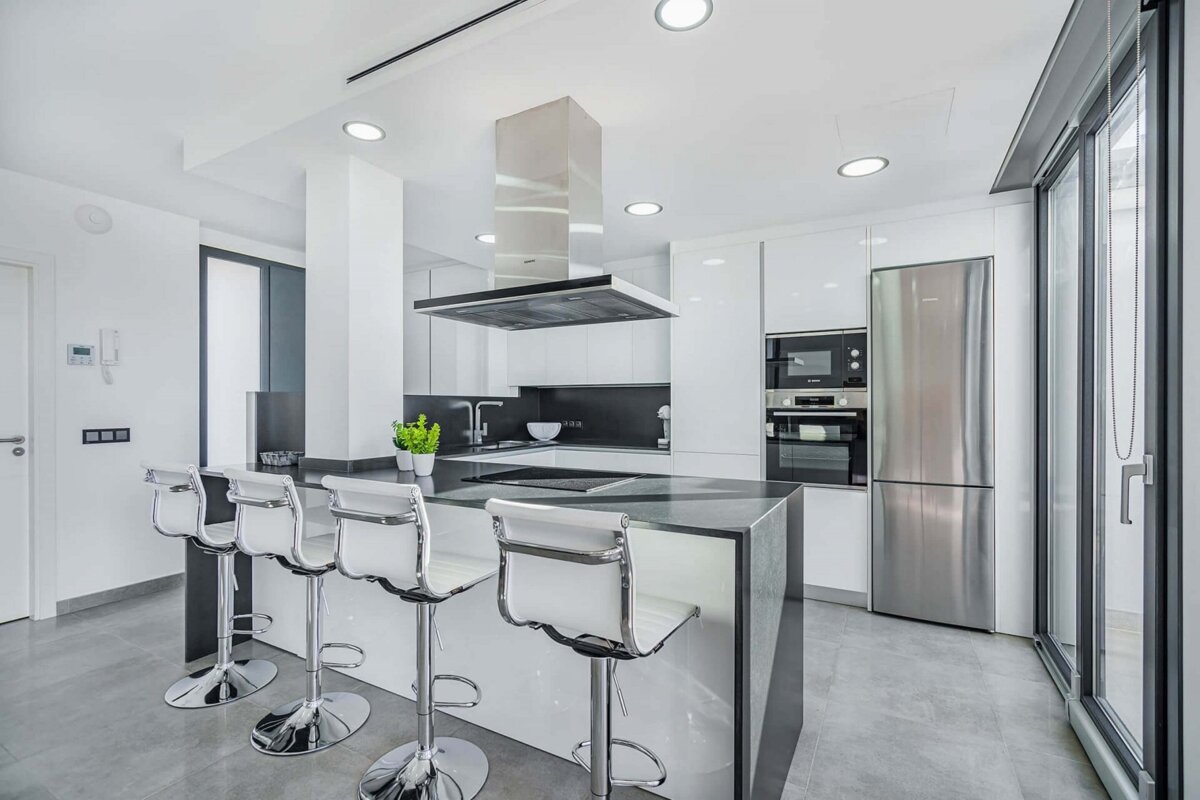 A sleek, modern white and grey kitchen features a large island with four bar stools, stainless steel appliances, and bright recessed lighting.