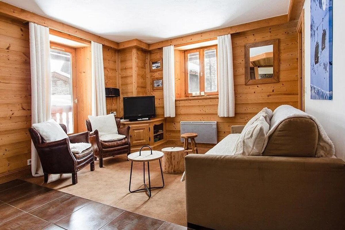 A warm, wood-paneled living room featuring a sofa, two leather armchairs, a TV, and windows with white curtains, exuding a rustic chalet vibe.
