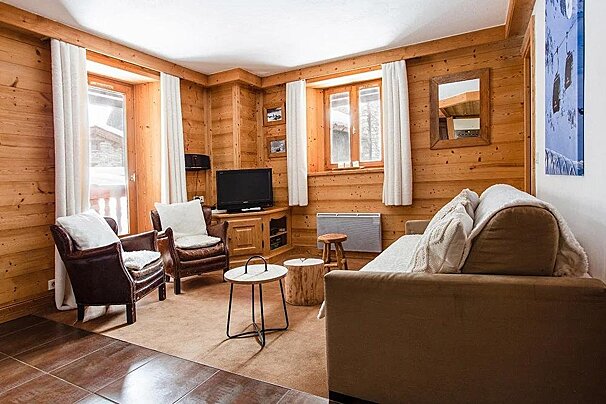 A warm, wood-paneled living room featuring a sofa, two leather armchairs, a TV, and windows with white curtains, exuding a rustic chalet vibe.