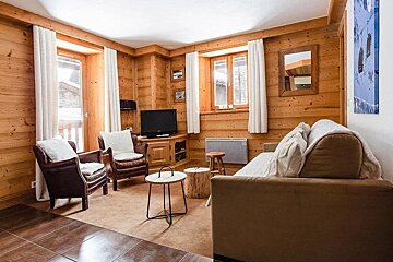 A warm, wood-paneled living room featuring a sofa, two leather armchairs, a TV, and windows with white curtains, exuding a rustic chalet vibe.