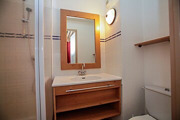 A compact bathroom features a shower, wooden vanity with sink and mirror, and toilet. White walls and blue accent tiles complete the look.