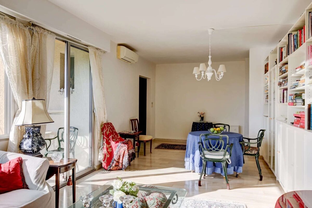 A bright, sunlit living/dining room features wood floors, sliding glass doors, a blue-draped dining table, green chairs, and a large bookshelf.