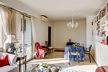 A bright, sunlit living/dining room features wood floors, sliding glass doors, a blue-draped dining table, green chairs, and a large bookshelf.