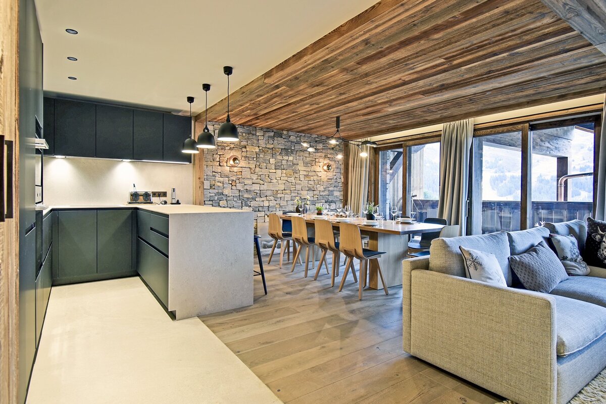 Modern chalet interior with a wooden ceiling, stone accent wall, dark kitchen cabinets, dining table, and cozy living area.