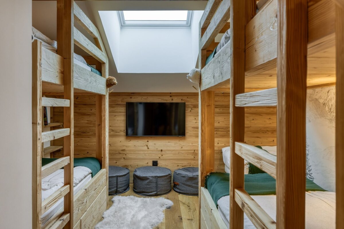 Cozy room with facing wooden bunk beds, a TV on wood-paneled walls, a skylight, grey poufs, and a white fluffy rug.