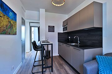 A modern kitchenette with gray cabinets, dark countertops, and a bar table with two stools. Wood flooring and a vibrant painting complete the sleek space.