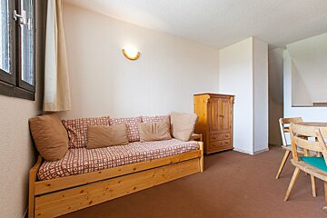 A cozy room with a wooden daybed, patterned cushions, a wooden wardrobe, and a dining table on brown carpet. Window left, wall light center.
