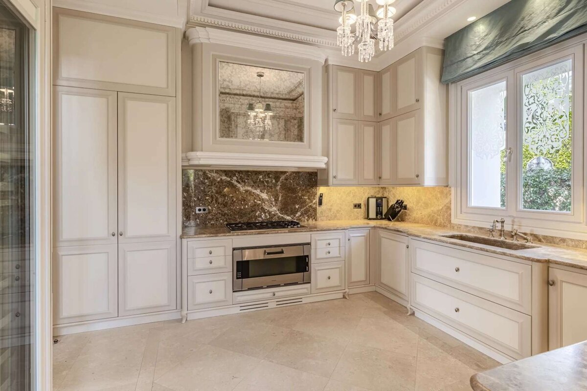 An elegant kitchen featuring light cabinets, luxurious marble countertops and backsplash, a crystal chandelier, and a large window with decorative glass.
