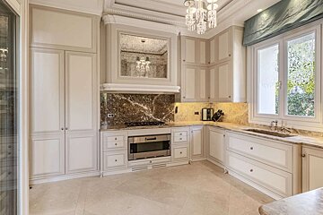 An elegant kitchen featuring light cabinets, luxurious marble countertops and backsplash, a crystal chandelier, and a large window with decorative glass.