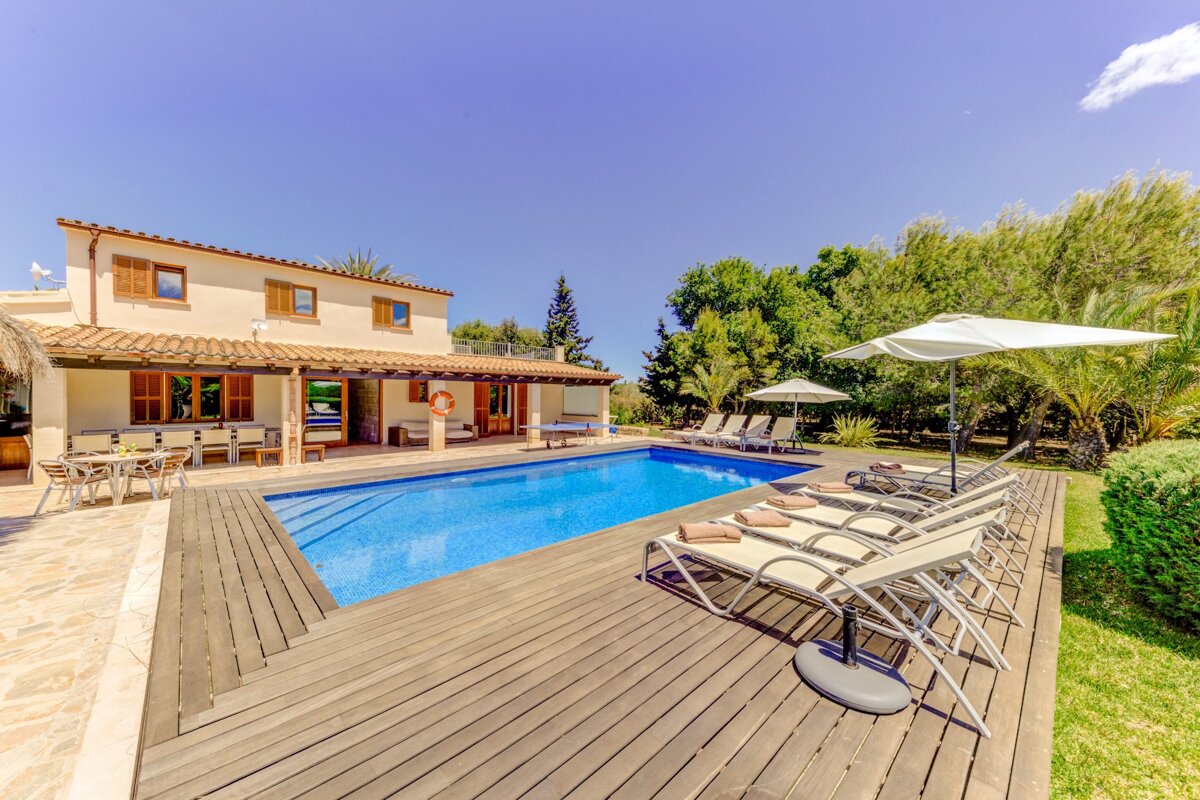 A beautiful villa with a sparkling blue swimming pool, wooden deck, sun loungers, and lush green trees under a clear sky.