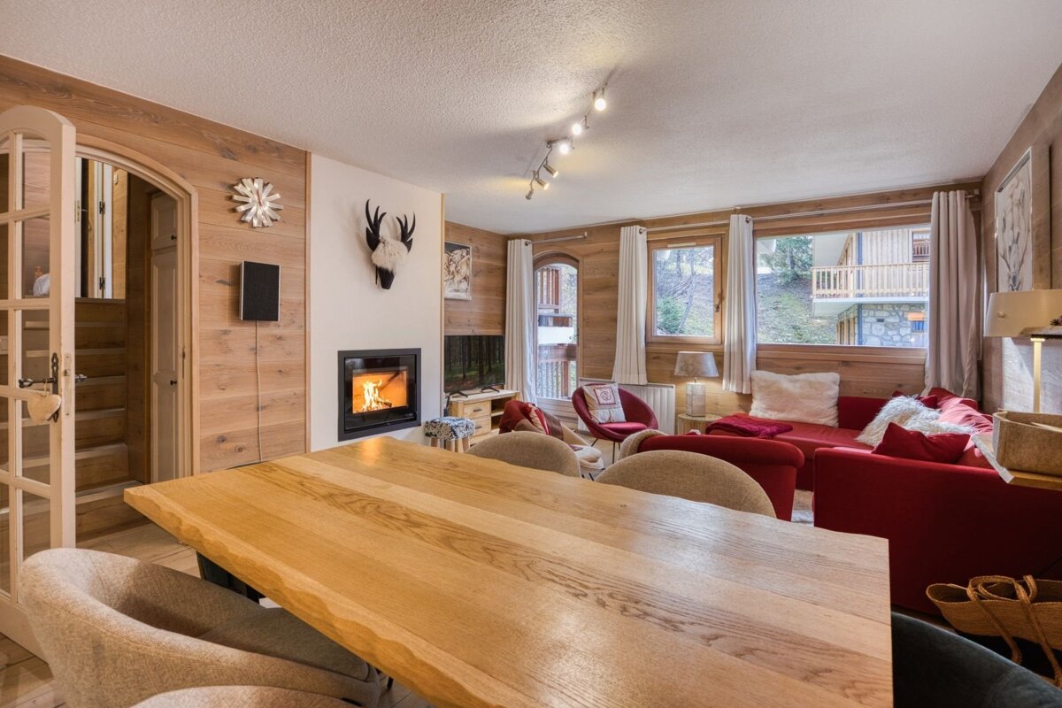 Inviting, wood-paneled chalet living space featuring a fireplace, red sofas, a large wooden dining table, and windows with outdoor views.