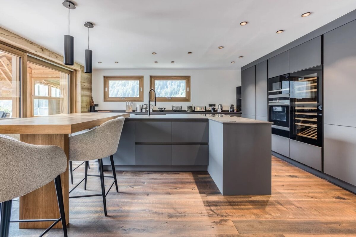 Stylish modern kitchen with dark gray cabinets, a wooden island bar with stools, and warm wood floors. Integrated appliances and scenic window views.