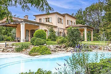 A charming peach-colored villa with terracotta roof and blue shutters, nestled in a lush garden with a sparkling blue pool and shaded patios.