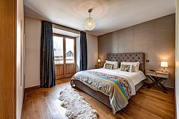 A modern rustic bedroom featuring a large bed with a tufted headboard, wooden floor, and French doors opening to an exterior view. A cozy rug and chandelier add warmth.
