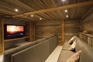 A cozy, rustic home theater room with wood-paneled walls and ceiling, recessed lighting, a large TV showing a sunset, and two long gray sofas.