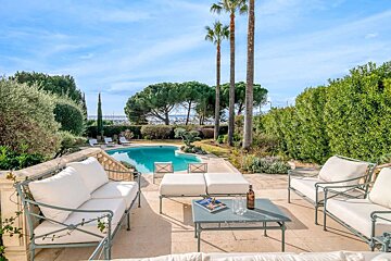 Elegant patio with white cushioned seating, a blue swimming pool, surrounded by lush greenery and palm trees, offering a scenic view of a distant marina.