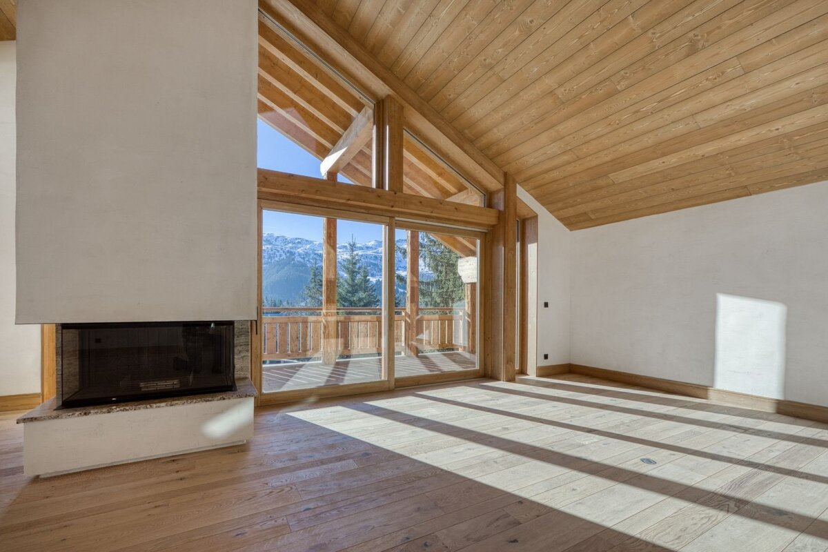 A sunny, empty room features light wood floors/ceiling, a modern fireplace, and large windows with a stunning mountain view and balcony.