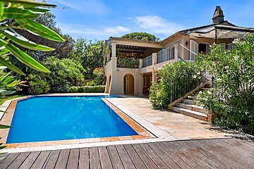 Elegant villa with a sparkling blue swimming pool, lush greenery, and paved patios under a bright sky.