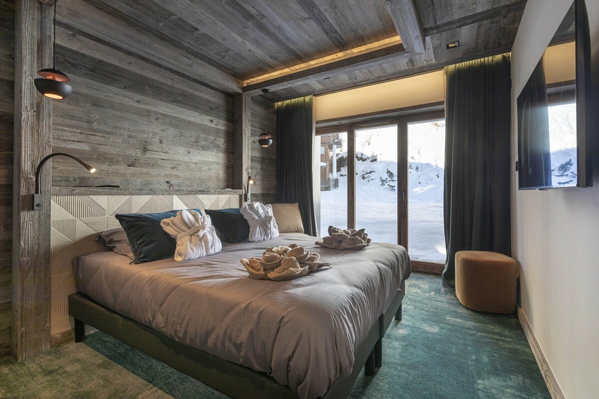 Cozy wooden bedroom with a large bed, robes, and a stunning snowy mountain view through floor-to-ceiling windows, featuring warm lighting.