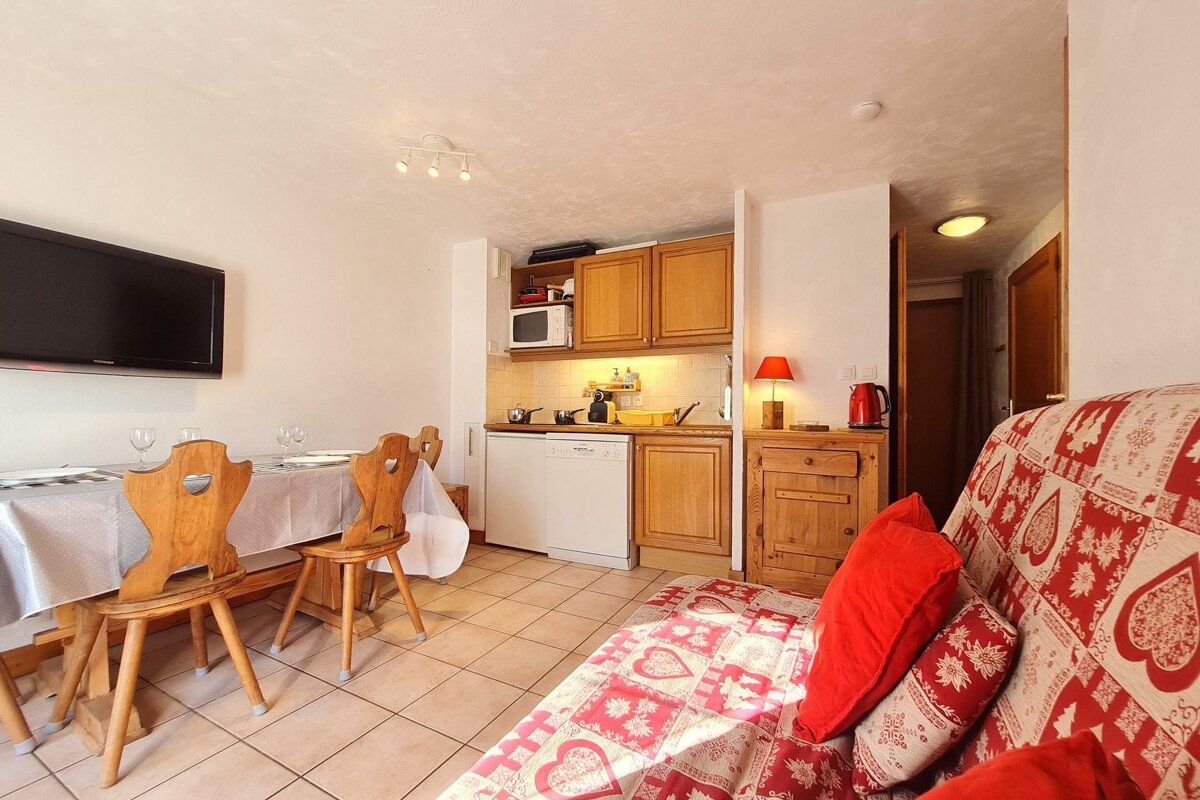 A bright, compact apartment with a kitchenette, dining area, wall-mounted TV, and a sofa featuring a red and white chalet pattern.