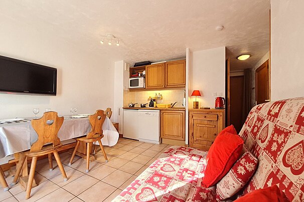 A bright, compact apartment with a kitchenette, dining area, wall-mounted TV, and a sofa featuring a red and white chalet pattern.