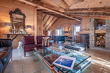 A cozy, wood-paneled chalet living room featuring a stone fireplace, bar, and comfortable seating, all bathed in warm sunlight.
