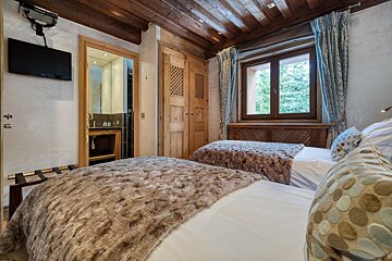 Cozy twin bedroom with rustic wooden details, fuzzy blankets, and a window view of trees. A bathroom and wall-mounted TV are also visible.