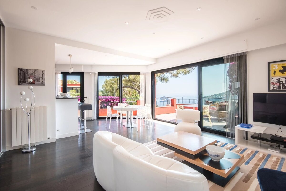Bright, modern living room with a white sofa, dark floors, and sliding glass doors opening to a scenic balcony with sea views.