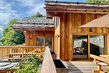 A beautiful wooden chalet with a stone roof and expansive wooden deck, featuring sliding glass doors, outdoor seating, and greenery under a sunny sky.