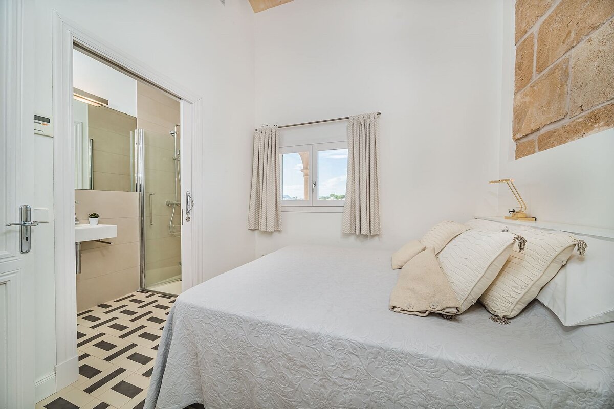 A bright white bedroom with a bed, window, and exposed brick wall. An open doorway reveals a modern bathroom with geometric floor tiles.